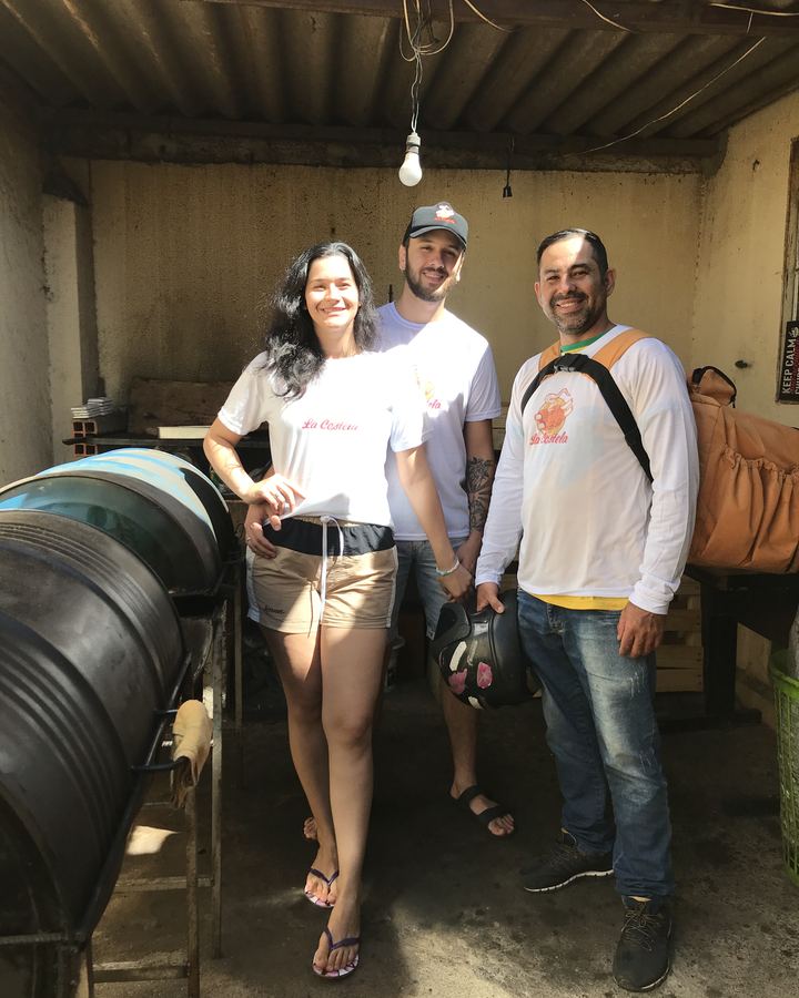 Três pessoas sorrindo no quintal, ao lado dos tambores de bafo. Uma delas segura um capacete e carrega a mochila de entrega.